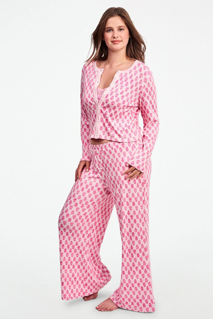 A woman stands against a white background, wearing the Daisy Paisley Nessa Cardigan by Roller Rabbit over a matching pink pajama set with red pineapple print, one hand on her hip.