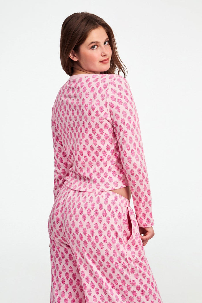 A woman stands against a white background, wearing the Daisy Paisley Nessa Cardigan by Roller Rabbit over a matching pink pajama set with red pineapple print, one hand on her hip.