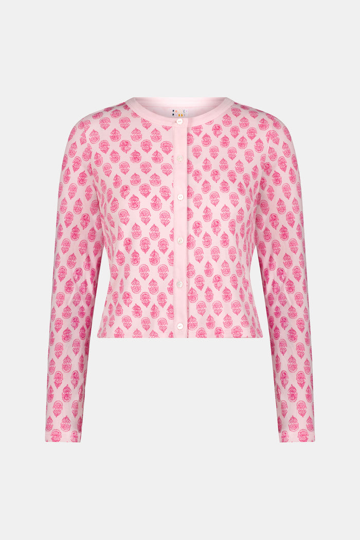 A woman stands against a white background, wearing the Daisy Paisley Nessa Cardigan by Roller Rabbit over a matching pink pajama set with red pineapple print, one hand on her hip.