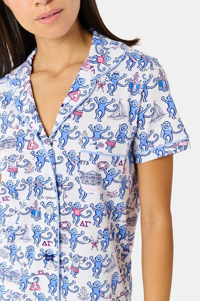 A woman in sunglasses and a white cap sits on outdoor steps, smiling and holding a cup of ice cream topped with colorful sprinkles. She is wearing the Roller Rabbit Delta Gamma Monkey Polo Pajamas—a printed short-sleeve romper with an elastic waistband. Another person holding ice cream is partially visible next to her.