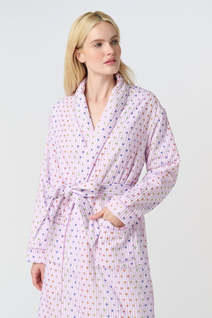 A person with long blonde hair stands barefoot against a plain white background, wearing the Roller Rabbit Disco Hearts Cosmo Robe with a belt tied at the waist.