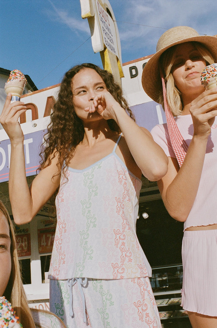 A woman stands barefoot wearing the Roller Rabbit Disco Monkey Lily Set, a matching Pima cotton pajama set featuring a pastel lace-like pattern, sleeveless camisole top, and drawstring pants with pockets.