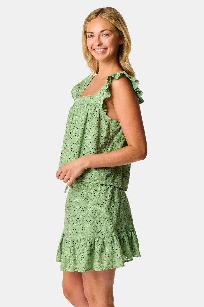 A person wearing the Roller Rabbit Elsa Eyelet Peyton Skirt in sage green, featuring a cotton eyelet fabric, ruffled hem, and elastic waistband, paired with a matching top and slightly lifting the top to reveal the skirt’s waist.
