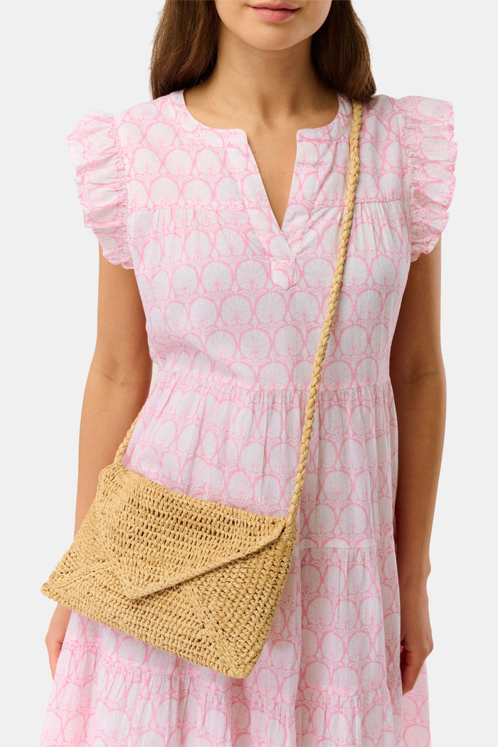 A woman in a light pink, short-sleeve, ruffled dress with a subtle pattern carries the Hat Attack Emmie Envelope Clutch, a woven beige raffia bag with flap closure. The image is cropped below her eyes.