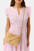 A woman in a light pink, short-sleeve, ruffled dress with a subtle pattern carries the Hat Attack Emmie Envelope Clutch, a woven beige raffia bag with flap closure. The image is cropped below her eyes.