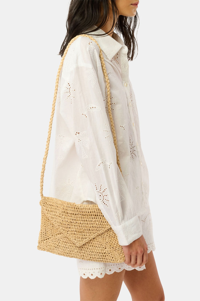A woman in a light pink, short-sleeve, ruffled dress with a subtle pattern carries the Hat Attack Emmie Envelope Clutch, a woven beige raffia bag with flap closure. The image is cropped below her eyes.