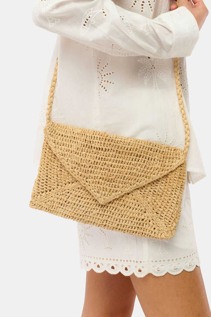A woman in a light pink, short-sleeve, ruffled dress with a subtle pattern carries the Hat Attack Emmie Envelope Clutch, a woven beige raffia bag with flap closure. The image is cropped below her eyes.