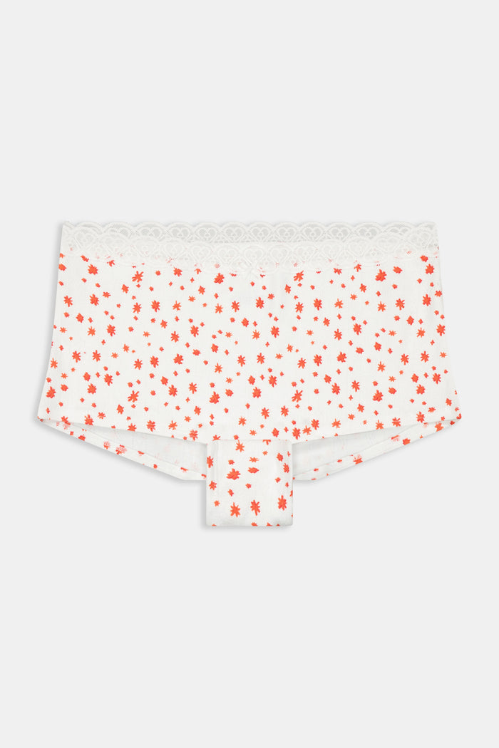 A person wears Roller Rabbit’s Etoiles Pointelle Crosby Boyshort—white shorts with lace trim and small red florals—paired with a light blue croissant-print camisole, standing against a plain white background.