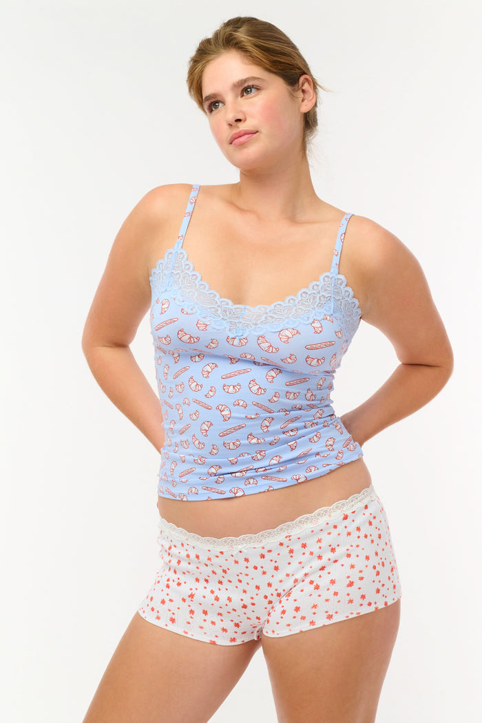 A person wears Roller Rabbit’s Etoiles Pointelle Crosby Boyshort—white shorts with lace trim and small red florals—paired with a light blue croissant-print camisole, standing against a plain white background.