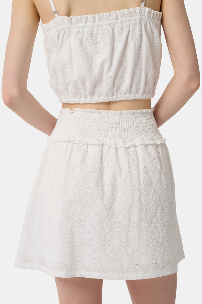 Wearing the Roller Rabbit Eyelet Lattice Erin Skirt with a smocked waist and delicate embroidery, paired with a matching white cropped top, their hand rests on their hip.