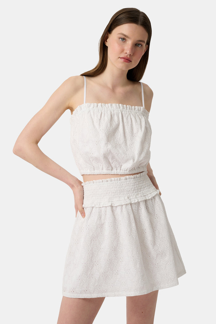 A woman wears the Roller Rabbit Eyelet Lattice Leah Top—white with thin straps and ruffled, elastic detail—paired with a matching high-waisted skirt. This stylish summer look is set against a plain white background.
