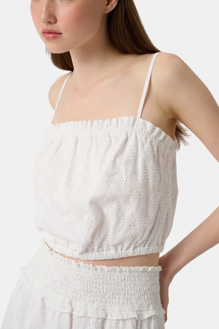 A woman wears the Roller Rabbit Eyelet Lattice Leah Top—white with thin straps and ruffled, elastic detail—paired with a matching high-waisted skirt. This stylish summer look is set against a plain white background.