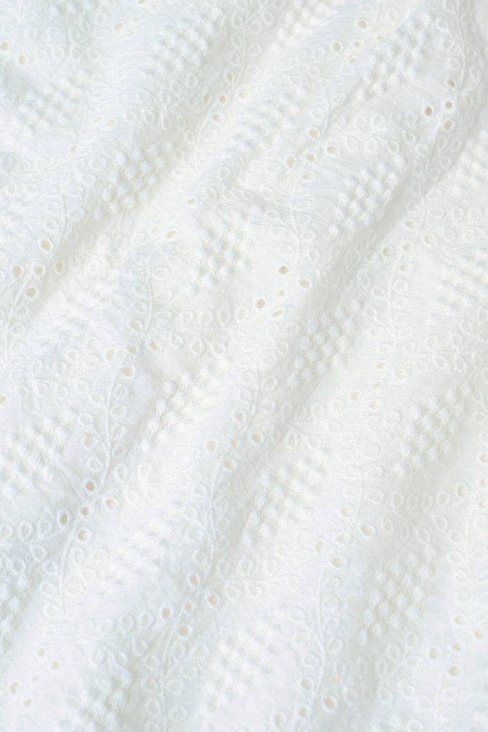A woman wears the Roller Rabbit Eyelet Lattice Leah Top—white with thin straps and ruffled, elastic detail—paired with a matching high-waisted skirt. This stylish summer look is set against a plain white background.