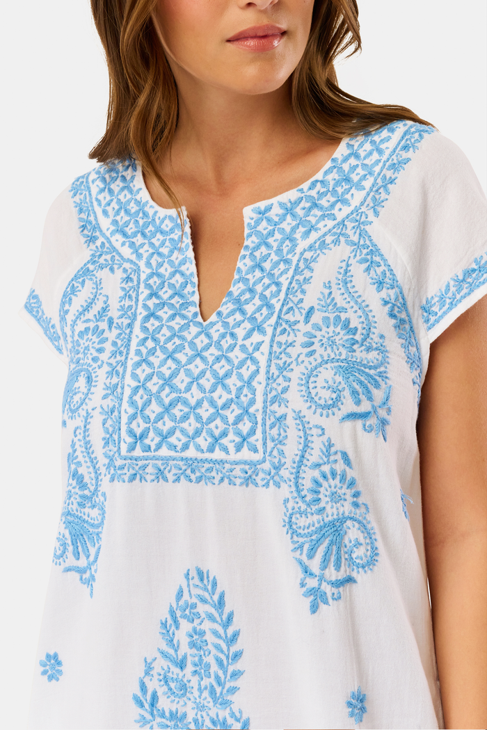 A woman wears the Roller Rabbit Faith Dress, a short-sleeved white cotton midi dress with blue embroidery, standing facing forward with hands by her sides against a plain light background.
