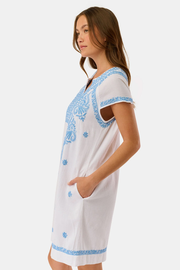 A woman wears the Roller Rabbit Faith Dress, a short-sleeved white cotton midi dress with blue embroidery, standing facing forward with hands by her sides against a plain light background.