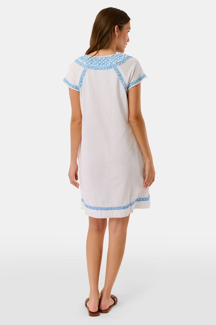 A woman wears the Roller Rabbit Faith Dress, a short-sleeved white cotton midi dress with blue embroidery, standing facing forward with hands by her sides against a plain light background.