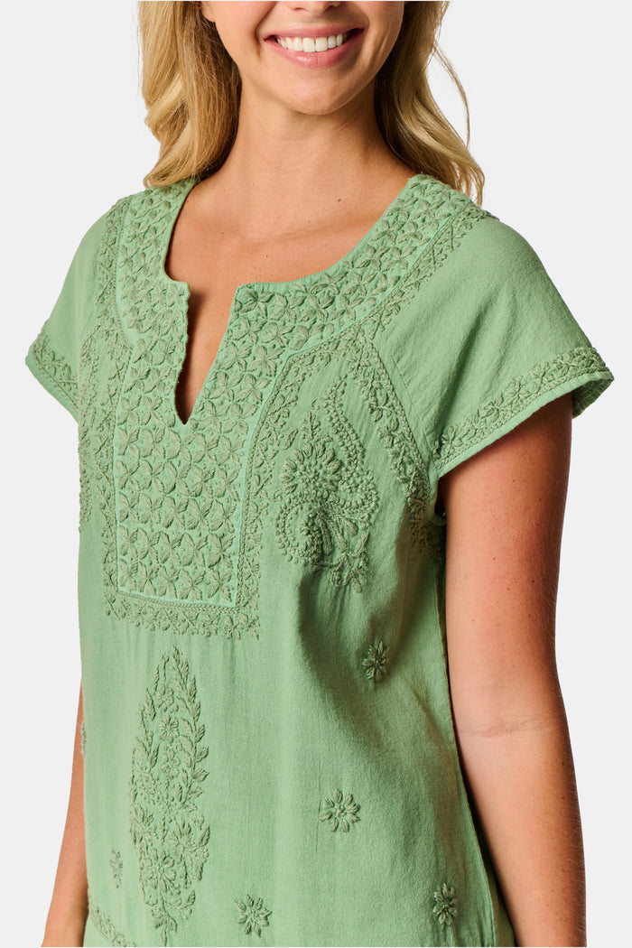 A woman wearing the Roller Rabbit Faith Top—a light green, short-sleeve embroidered cotton blouse with a split neckline—paired with white pants, stands against a plain light background.
