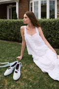 A woman wearing the Roller Rabbit Florence Lace Mae Top sits on the grass near hedges, looking to the side. Light blue ballet flats with ribbons lie beside her, with a house featuring large windows in the background.