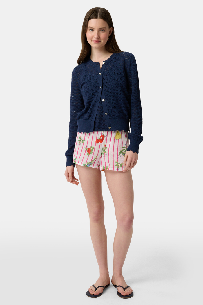 A person stands hands on hips, wearing a navy blue cardigan and Roller Rabbit Fruits D'Ete Sonny Boxer in white cotton voile, featuring pink stripes plus colorful strawberries, oranges, pears, and floral patterns.