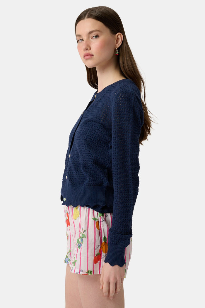 A person stands hands on hips, wearing a navy blue cardigan and Roller Rabbit Fruits D'Ete Sonny Boxer in white cotton voile, featuring pink stripes plus colorful strawberries, oranges, pears, and floral patterns.