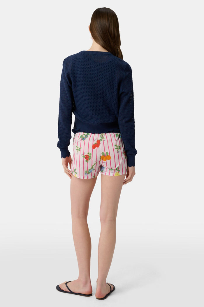 A person stands hands on hips, wearing a navy blue cardigan and Roller Rabbit Fruits D'Ete Sonny Boxer in white cotton voile, featuring pink stripes plus colorful strawberries, oranges, pears, and floral patterns.