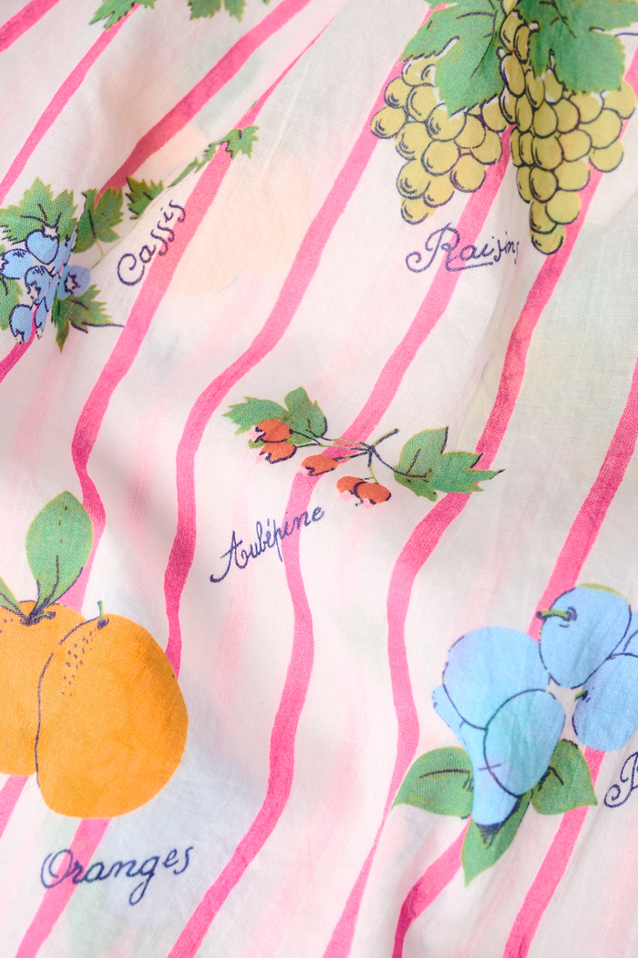 A woman in the Roller Rabbit Fruits D'Ete Keaton Dress with a matching headscarf stands barefoot by a vintage white Volkswagen van, shielding her eyes from the sun. The sunny coastal backdrop highlights the dress's playful print.