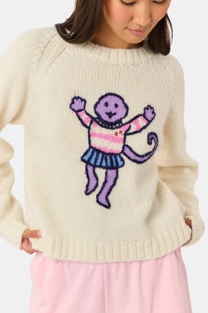 A young woman sits on a bench, wearing the Roller Rabbit Game Day Brittany Sweater with a cartoon figure, layered over a light dress. She is beside a blue patterned backpack and surrounded by greenery.