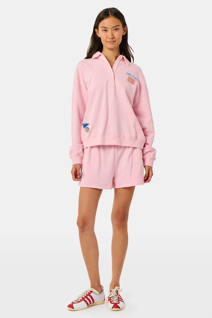 A young woman in a pink sweatshirt and Roller Rabbit Game Day Nicki Shorts smiles while holding a football in one hand, standing on a grassy field with trees and buildings in the background.