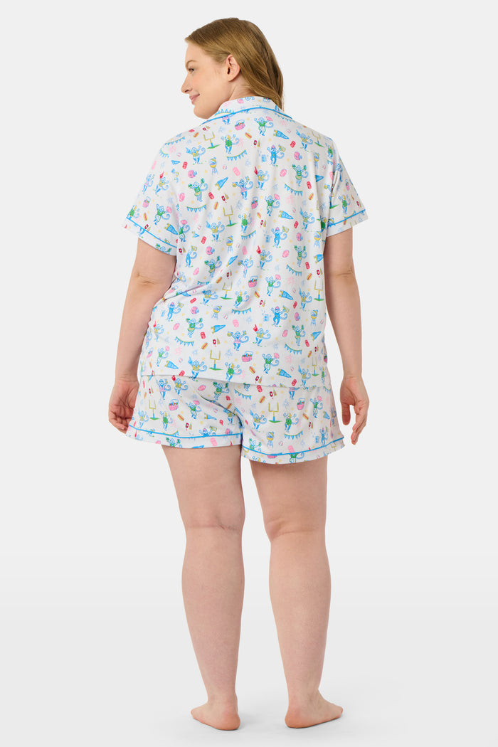 The Game Day Polo Pajamas by Roller Rabbit feature a short-sleeve button-up top and shorts, adorned with a lively pattern of colorful cartoon animals and party items on a white background.