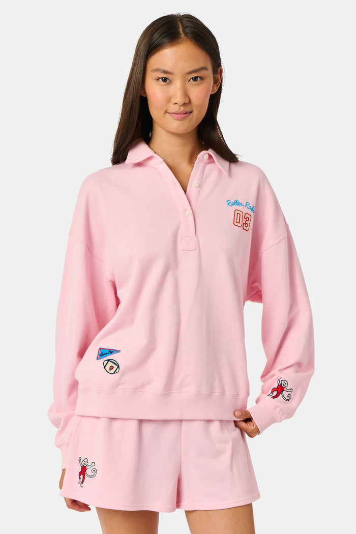 A young woman in the Roller Rabbit Game Day Rugby Sweatshirt and cap smiles while holding a football on a grassy field with trees and buildings in the background.