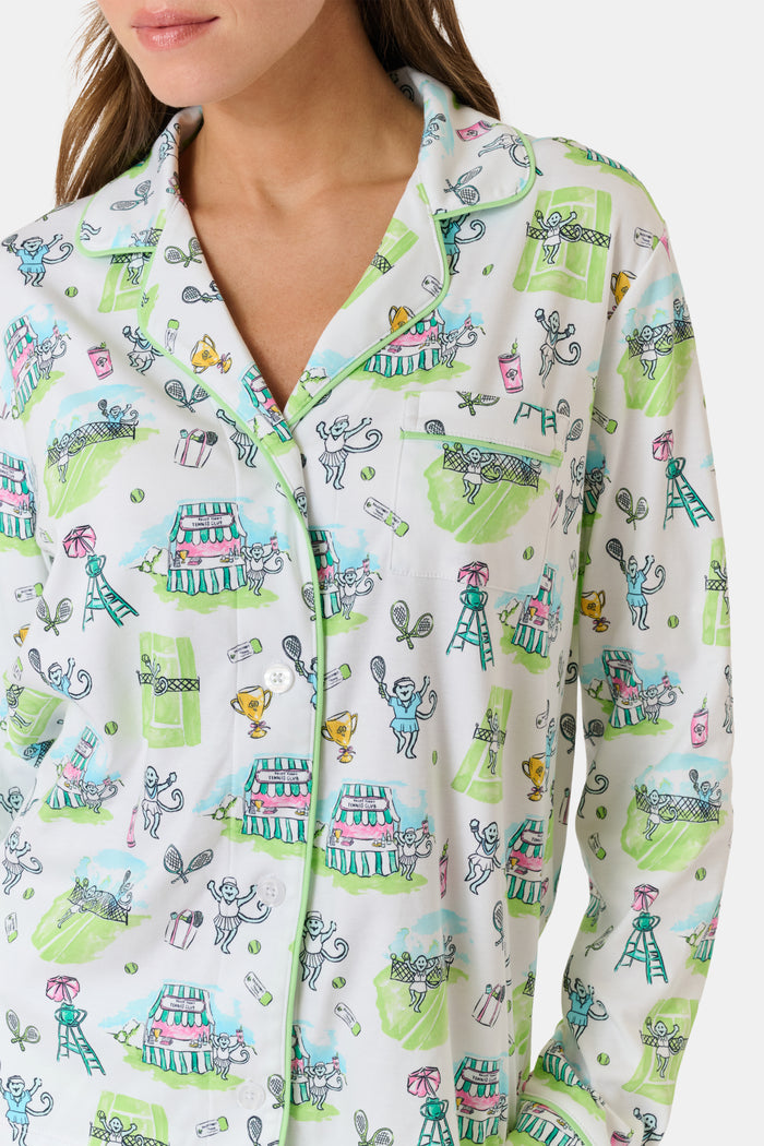Introducing the Game, Set, Monkey Long Sleeve Polo Pajamas by Roller Rabbit—a cozy white pajama set expertly crafted from soft Pima Cotton. The set features a playful Game Set Monkey print with vibrant green, blue, and yellow animal and nature illustrations. Designed with long sleeves, a button-up polo top, and comfortable drawstring waist pants, this delightful set is showcased against a plain light background.