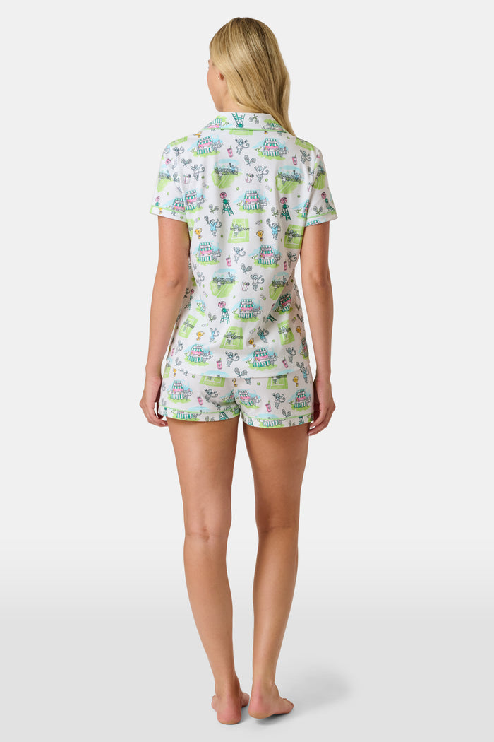 The Roller Rabbit Game, Set, Monkey Polo Pajamas is a two-piece set featuring a short-sleeve button-up shirt and matching shorts. Crafted from soft Pima Cotton, the white fabric is adorned with a playful cartoon animal and house print in green, blue, and yellow tones.