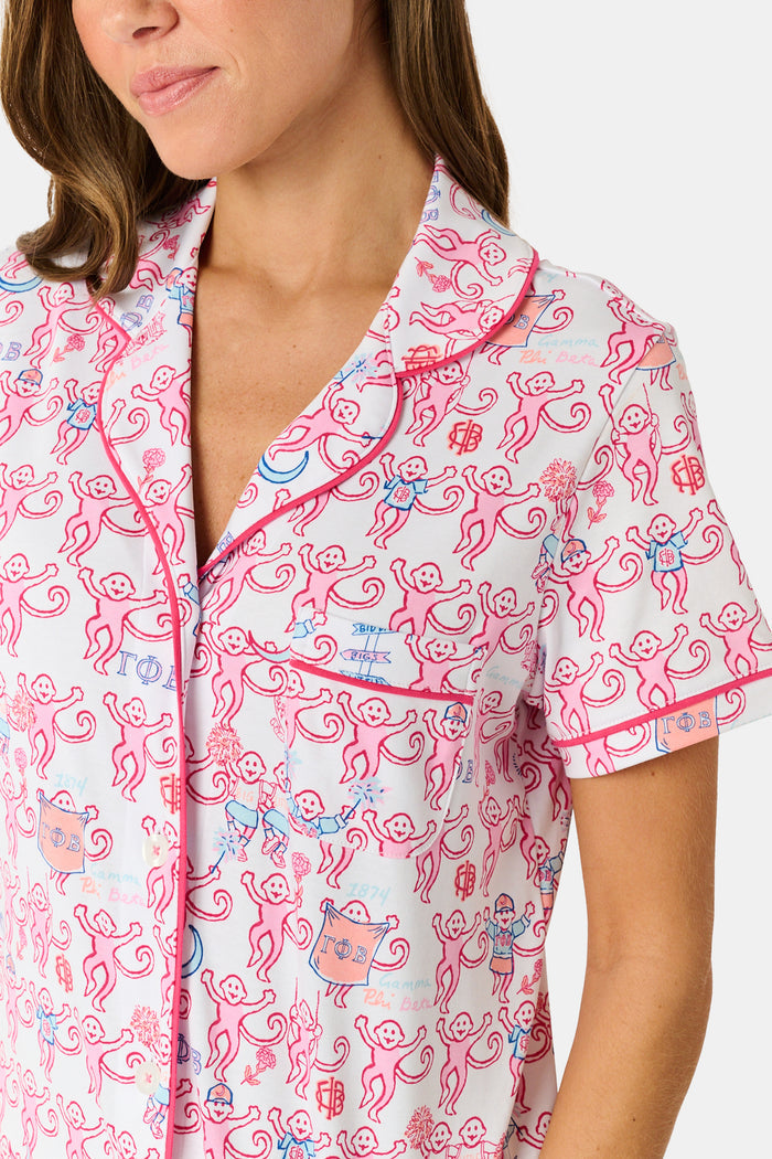Introducing the Roller Rabbit Gamma Phi Beta Monkey Polo Pajamas: a short-sleeve Pima cotton set featuring a button-up top and matching shorts, adorned with a whimsical pink and blue monkey pattern and red trim on a crisp white background.
