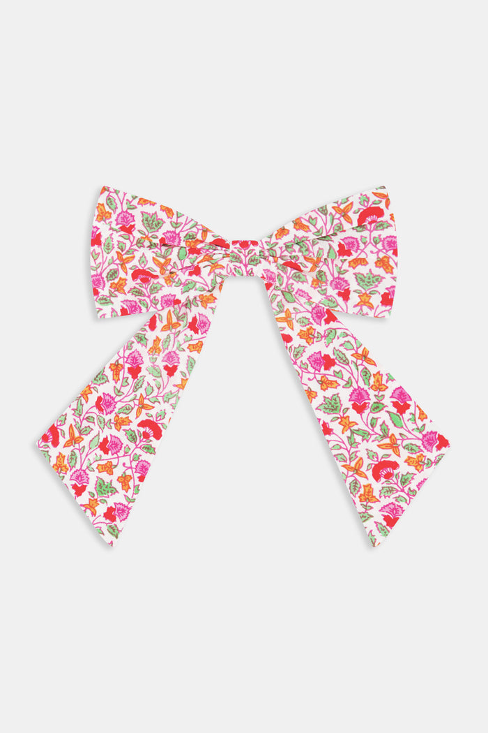 The Garden Gate Floral Hair Bow by Roller Rabbit is a large cotton bow with long tails, showcasing a vibrant pattern of pink, red, and orange flowers with green leaves on a white background.