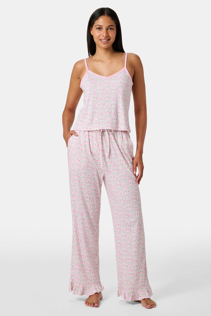 The Garden Gate Floral Lily Set by Roller Rabbit features a pink and white women's loungewear set with a sleeveless, V-neck camisole and loose pants. Made from soft Pima cotton, both pieces showcase a delicate floral pattern and light pink trim. The comfy tank and pants include a drawstring waist and ruffled hems for added comfort and style.