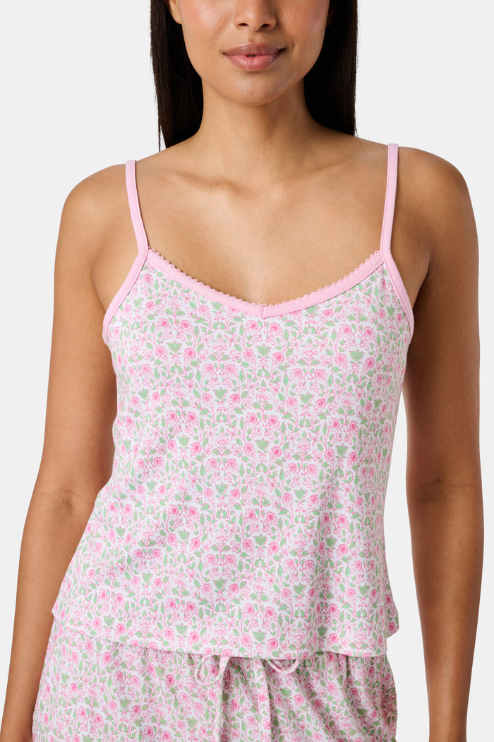 The Garden Gate Floral Lily Set by Roller Rabbit features a pink and white women's loungewear set with a sleeveless, V-neck camisole and loose pants. Made from soft Pima cotton, both pieces showcase a delicate floral pattern and light pink trim. The comfy tank and pants include a drawstring waist and ruffled hems for added comfort and style.