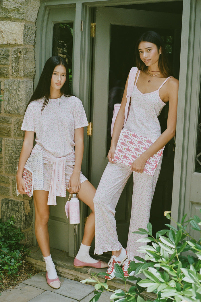 The Garden Gate Floral Lily Set by Roller Rabbit features a pink and white women's loungewear set with a sleeveless, V-neck camisole and loose pants. Made from soft Pima cotton, both pieces showcase a delicate floral pattern and light pink trim. The comfy tank and pants include a drawstring waist and ruffled hems for added comfort and style.