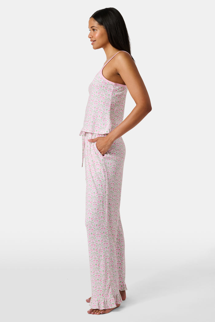 The Garden Gate Floral Lily Set by Roller Rabbit features a pink and white women's loungewear set with a sleeveless, V-neck camisole and loose pants. Made from soft Pima cotton, both pieces showcase a delicate floral pattern and light pink trim. The comfy tank and pants include a drawstring waist and ruffled hems for added comfort and style.