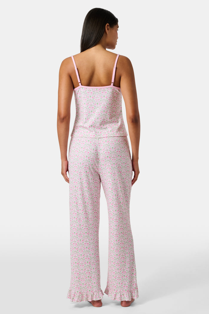 The Garden Gate Floral Lily Set by Roller Rabbit features a pink and white women's loungewear set with a sleeveless, V-neck camisole and loose pants. Made from soft Pima cotton, both pieces showcase a delicate floral pattern and light pink trim. The comfy tank and pants include a drawstring waist and ruffled hems for added comfort and style.