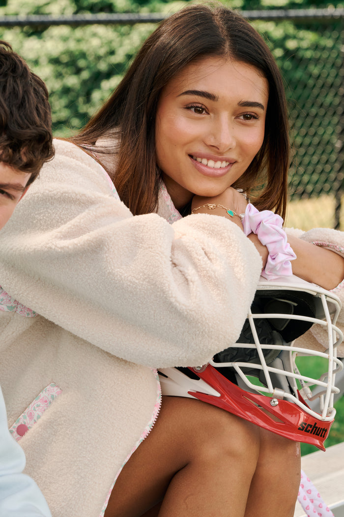 A young woman with long hair and a Roller Rabbit Garden Gate Floral Marina Fleece Jacket smiles while sitting outdoors, resting her arms on her knees next to a white and red helmet, with greenery and a fence in the background.