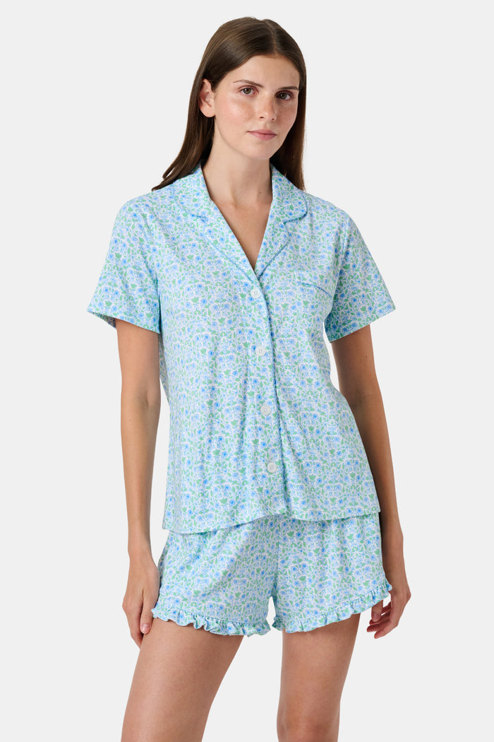 The Roller Rabbit Garden Gate Floral Wyatt Polo Pajamas feature a light blue short-sleeve set crafted from soft Pima Cotton with a delicate Garden Gate Floral Ditsy pattern. The button-up top is designed with a collar and chest pocket, while the matching shorts are accented with a charming ruffled hem. The background is plain white.