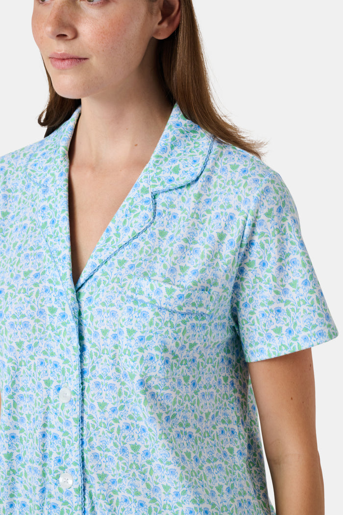 The Roller Rabbit Garden Gate Floral Wyatt Polo Pajamas feature a light blue short-sleeve set crafted from soft Pima Cotton with a delicate Garden Gate Floral Ditsy pattern. The button-up top is designed with a collar and chest pocket, while the matching shorts are accented with a charming ruffled hem. The background is plain white.