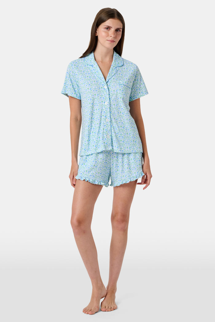 The Roller Rabbit Garden Gate Floral Wyatt Polo Pajamas feature a light blue short-sleeve set crafted from soft Pima Cotton with a delicate Garden Gate Floral Ditsy pattern. The button-up top is designed with a collar and chest pocket, while the matching shorts are accented with a charming ruffled hem. The background is plain white.