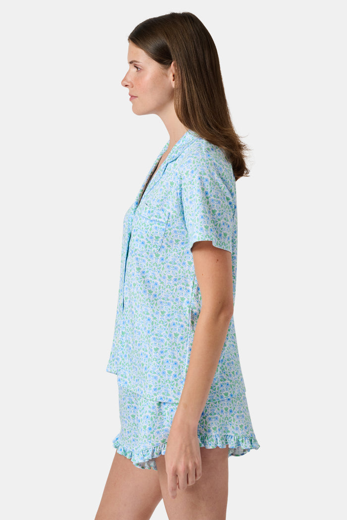 The Roller Rabbit Garden Gate Floral Wyatt Polo Pajamas feature a light blue short-sleeve set crafted from soft Pima Cotton with a delicate Garden Gate Floral Ditsy pattern. The button-up top is designed with a collar and chest pocket, while the matching shorts are accented with a charming ruffled hem. The background is plain white.