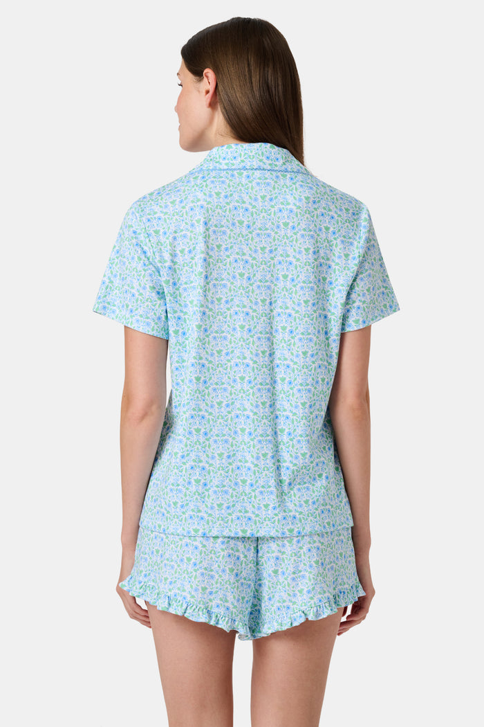 The Roller Rabbit Garden Gate Floral Wyatt Polo Pajamas feature a light blue short-sleeve set crafted from soft Pima Cotton with a delicate Garden Gate Floral Ditsy pattern. The button-up top is designed with a collar and chest pocket, while the matching shorts are accented with a charming ruffled hem. The background is plain white.
