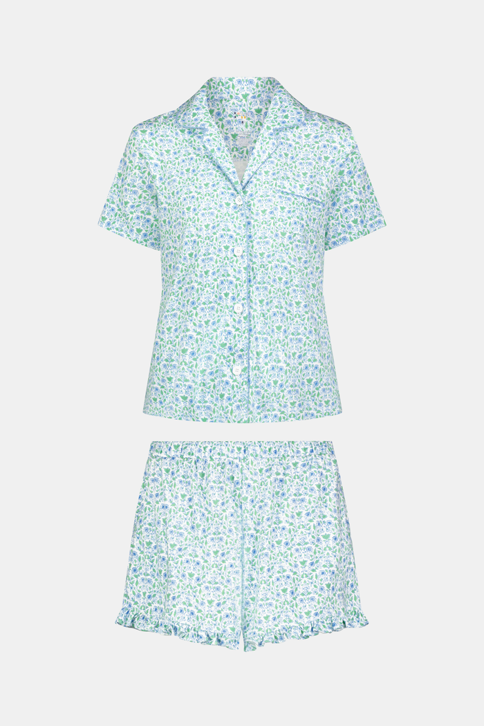 The Roller Rabbit Garden Gate Floral Wyatt Polo Pajamas feature a light blue short-sleeve set crafted from soft Pima Cotton with a delicate Garden Gate Floral Ditsy pattern. The button-up top is designed with a collar and chest pocket, while the matching shorts are accented with a charming ruffled hem. The background is plain white.