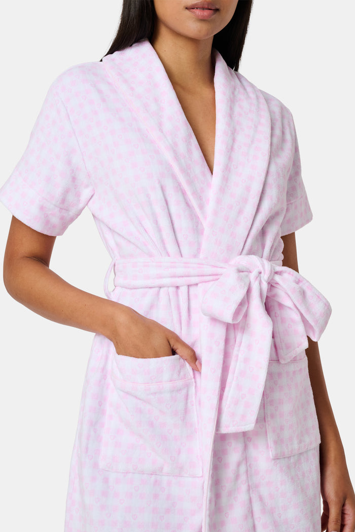 The Roller Rabbit Gingham Hearts Brooks Robe features a light pink gingham pattern with subtle hearts, crafted from soft cotton. This short-sleeve robe includes front pockets and a matching waist tie, and is displayed against a plain white background.