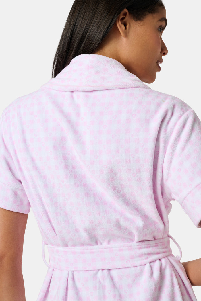 The Roller Rabbit Gingham Hearts Brooks Robe features a light pink gingham pattern with subtle hearts, crafted from soft cotton. This short-sleeve robe includes front pockets and a matching waist tie, and is displayed against a plain white background.