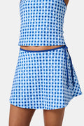 Wearing the Roller Rabbit Gingham Hearts Coco Wrap Skirt, a woman stands next to a net outdoors, surrounded by green grass and trees.