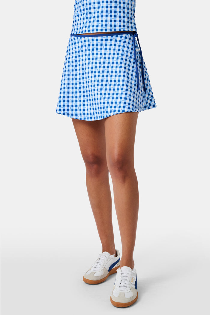 Wearing the Roller Rabbit Gingham Hearts Coco Wrap Skirt, a woman stands next to a net outdoors, surrounded by green grass and trees.